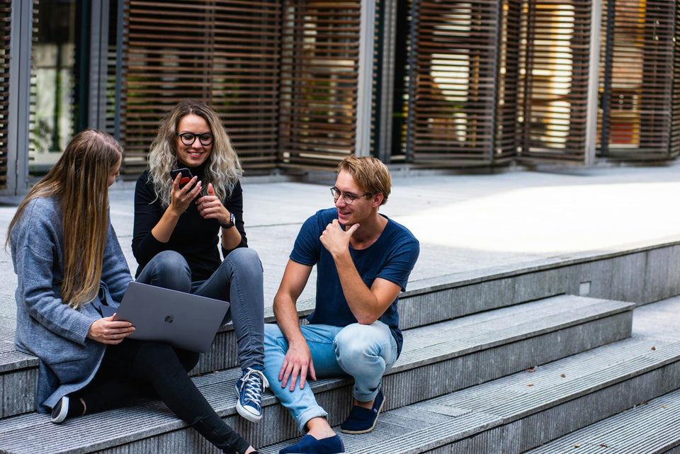 Drei Studierende sitzen mit Laptop und Smartphone auf Treppenstufen vor einem Universitätsgebäude