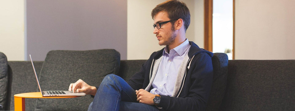 Junger Mann mit Brille und Hemd arbeitet konzentriert am Laptop auf einem Sofa