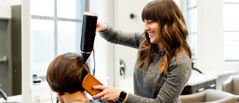 Eine lächelnde Friseurin föhnt und bürstet die Haare einer Kundin im Salon