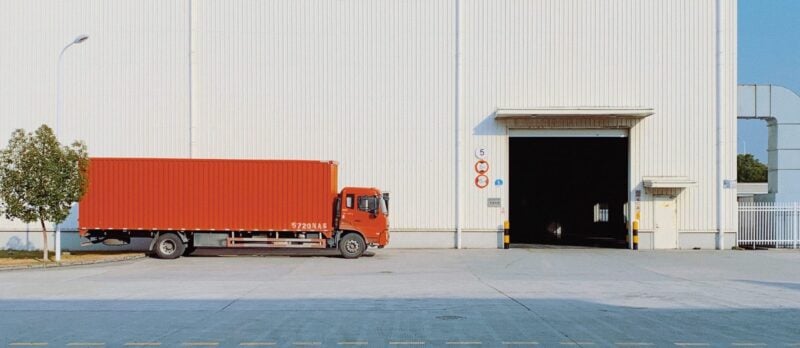 Ein roter Lastkraftwagen parkt vor dem offenen Tor einer großen, weißen Lagerhalle