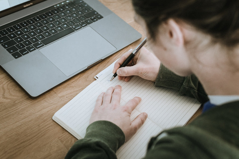 Eine Person schreibt konzentriert in ein Notizbuch neben einem Laptop zum Lernen für den Einstellungstest