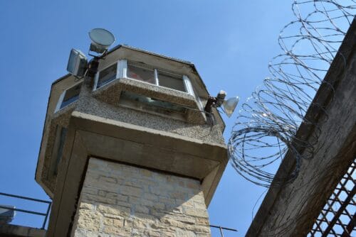 Wachturm einer Justizvollzugsanstalt mit Stacheldrahtzaun vor blauem Himmel zur Veranschaulichung der Arbeit im JVA-Dienst
