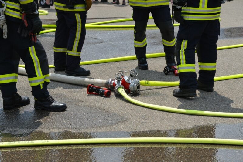 Feuerwehrleute in Schutzkleidung bei einer Übung mit Schläuchen und einem Verteiler auf nassem Asphalt